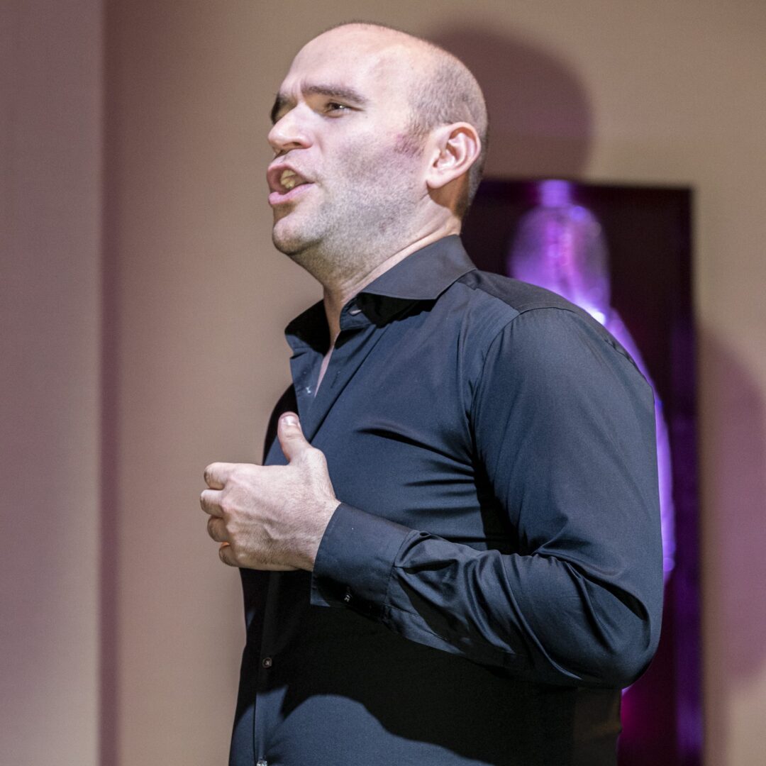 A bald man in a black shirt standing in front of a purple wall.